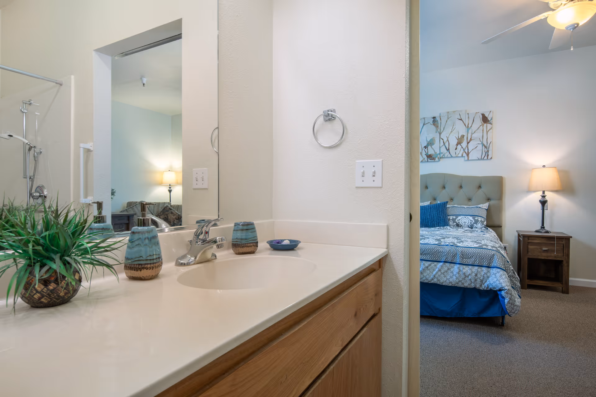 View of a bathroom sink countertop with a faucet, decorative plant, and small containers. A mirror reflects part of the bathroom, including a shower area. Through an open doorway, a bedroom is visible with a bed, nightstand, lamp, and wall art featuring birds on branches.
