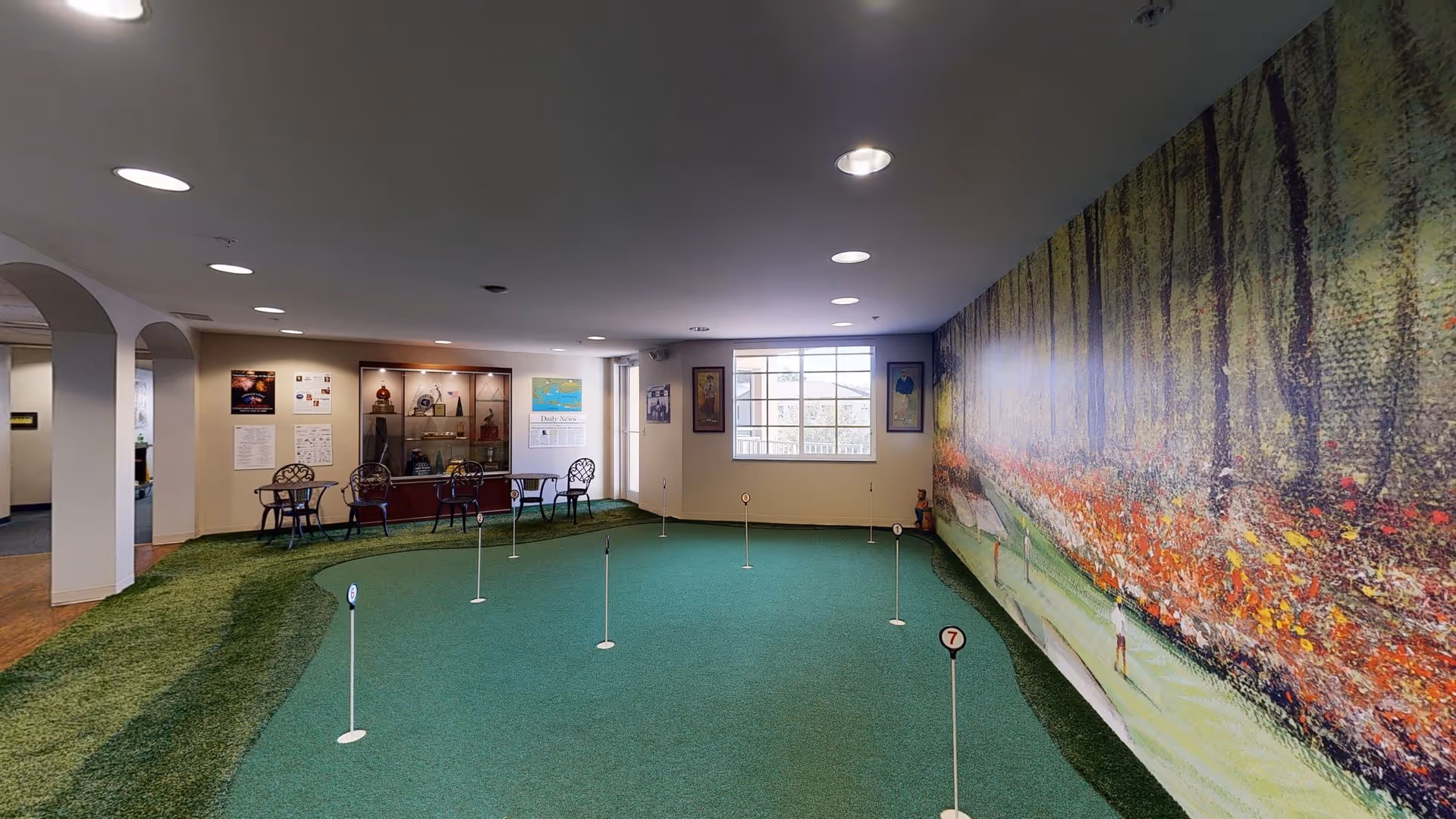 Indoor putting green area with several small golf holes marked by numbered flags. The room has a large mural on one wall depicting a golf course scene with trees and spectators. There are a few small tables and chairs along the far wall, and a display cabinet with trophies and awards. The space is well-lit with recessed ceiling lights and a window letting in natural light.