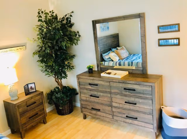 A bedroom corner featuring a wooden dresser with six drawers and a large mirror above it reflecting a bed with striped bedding and pillows. To the left of the dresser is a small wooden nightstand with two drawers, a lamp, and a framed photo. A tall potted plant is placed between the dresser and nightstand. On the right side, there is a white and blue laundry basket on the floor. The room has light-colored wooden flooring and white walls with two small framed pictures hanging beside the mirror.