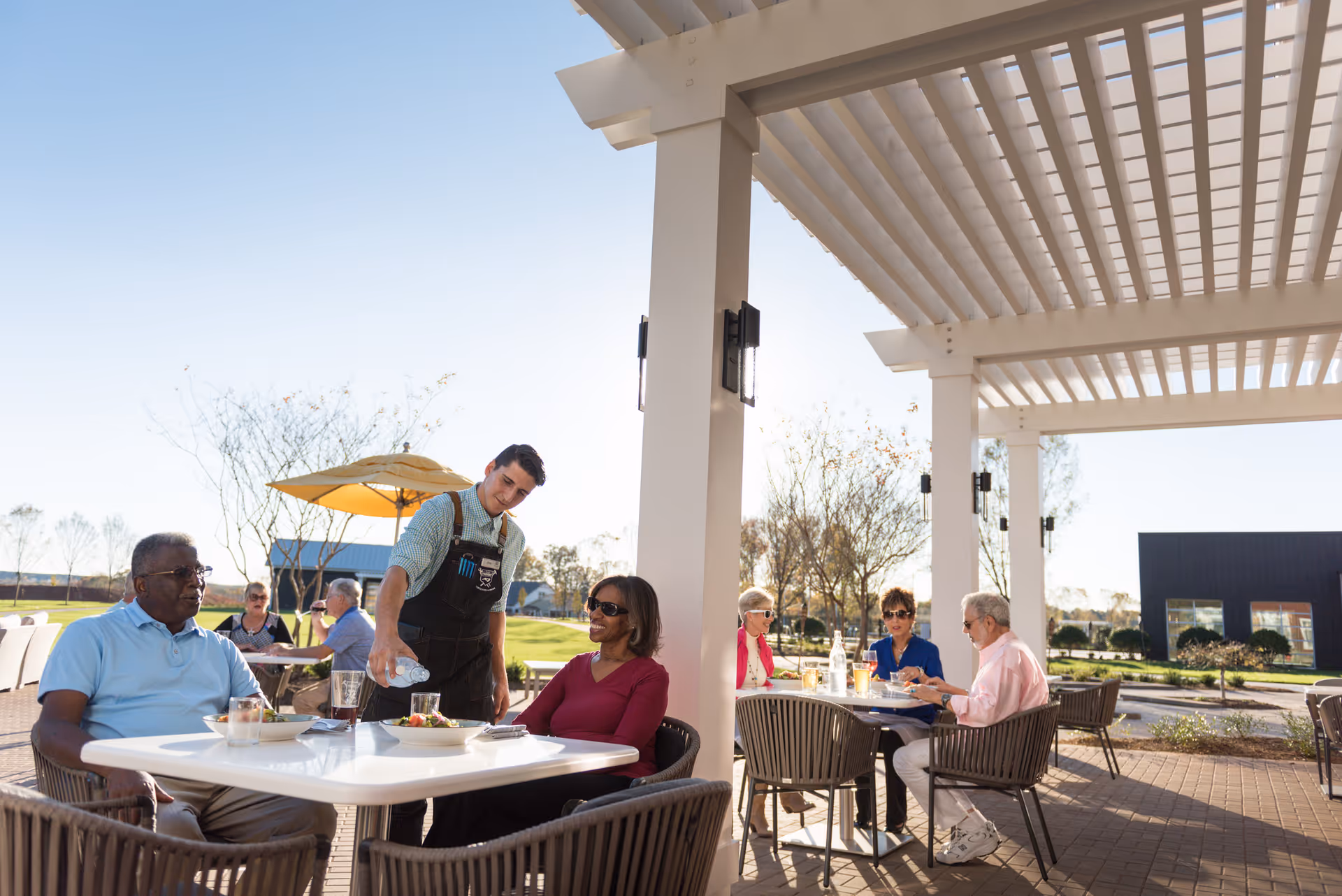 A group of seniors sitting at outdoor tables under a pergola at a resort community. A waiter is pouring water for a woman seated at one table. Other seniors are engaged in conversation at another table. The setting is bright and sunny with green grass and trees in the background.