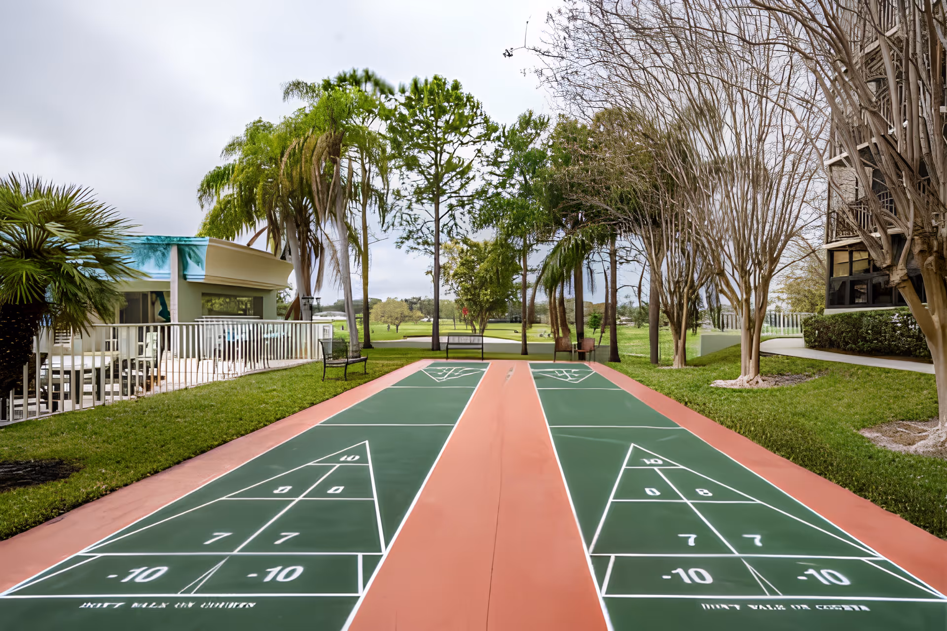 Two outdoor shuffleboard courts with green playing surfaces flanked by palm trees and nearby buildings.
