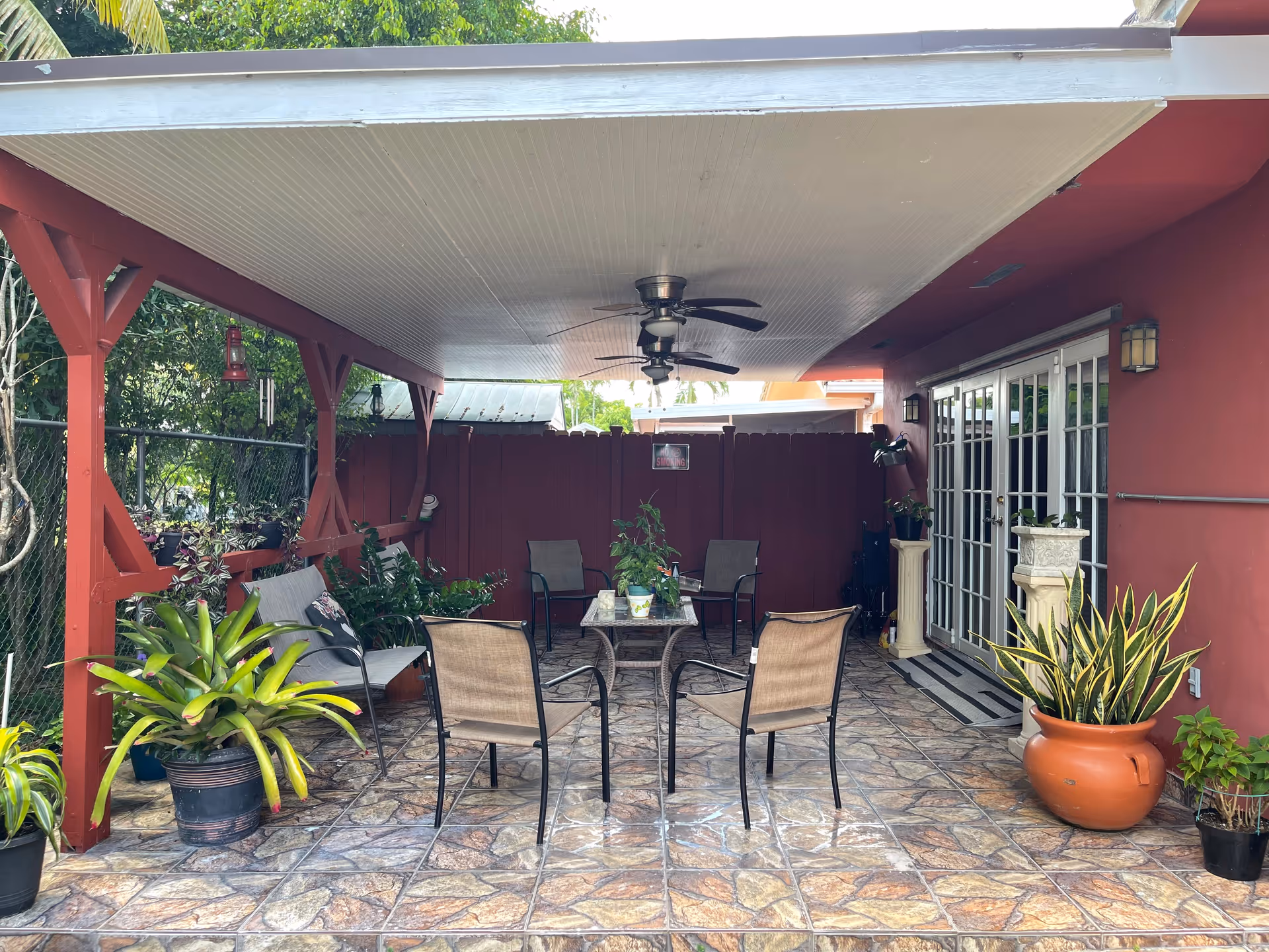 Covered outdoor patio area with a tiled floor, several chairs around a glass-top table, potted plants, ceiling fans, and a red wooden fence in the background. The patio is adjacent to a building with red walls and glass doors.