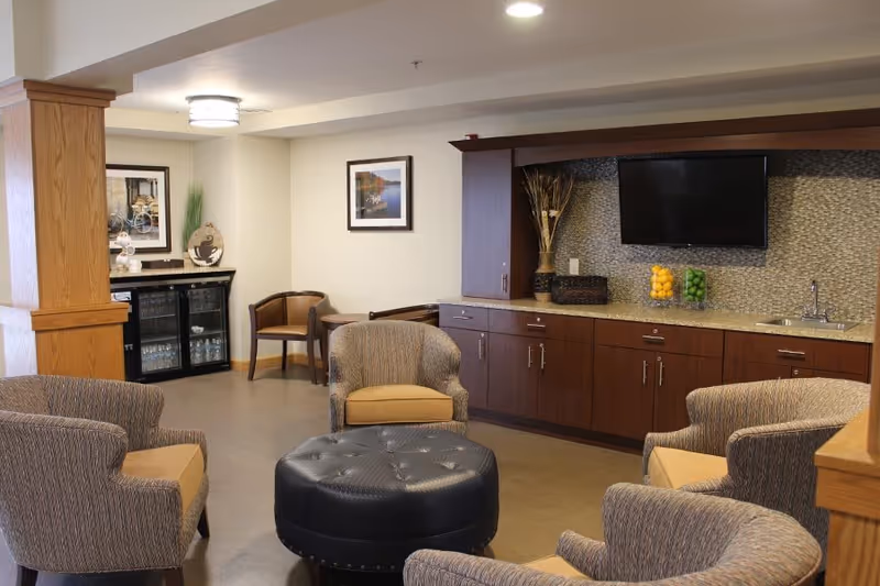 A cozy common area with four upholstered armchairs arranged around a round black ottoman. The room features wooden cabinetry with a granite countertop, a mounted flat-screen TV, and decorative items including vases with lemons and limes. There are framed pictures on the walls and a small table with two chairs in the corner.
