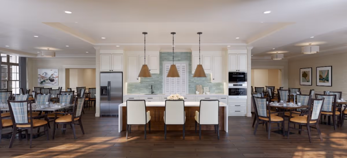 A spacious and well-lit dining room with multiple wooden tables and chairs arranged neatly. In the center, there is a kitchen island with three white cushioned chairs and three pendant lights hanging above. The kitchen area features white cabinetry, a stainless steel refrigerator, built-in ovens, and a light green tiled backsplash. The room has large windows allowing natural light to fill the space, and framed artwork is visible on the walls.