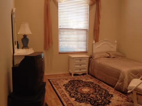 A small bedroom with a single bed covered in a beige quilt, a white nightstand with drawers, a window with blinds and curtains, a table lamp on a dresser, and an old-style CRT television on a stand. There is a patterned rug on the wooden floor.