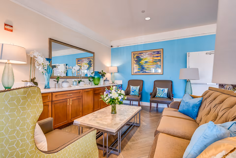 A bright and cozy living room in a senior living facility featuring a tan sofa with blue and beige pillows, two brown chairs with decorative cushions, a green patterned armchair, and a rectangular coffee table with a flower arrangement. The room has a blue accent wall with a colorful framed painting, a large mirror above a wooden cabinet with various vases and plants, and several table lamps providing warm lighting.