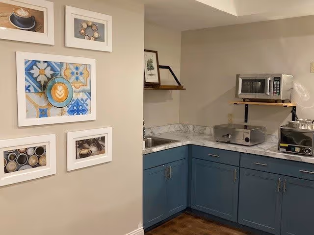 A small kitchen area with blue lower cabinets and white marble countertops. On the right side, there is a microwave on a wooden shelf, a toaster oven, and a coffee maker. On the left wall, there are four framed pictures featuring coffee cups and coffee-related themes. The walls are painted beige.