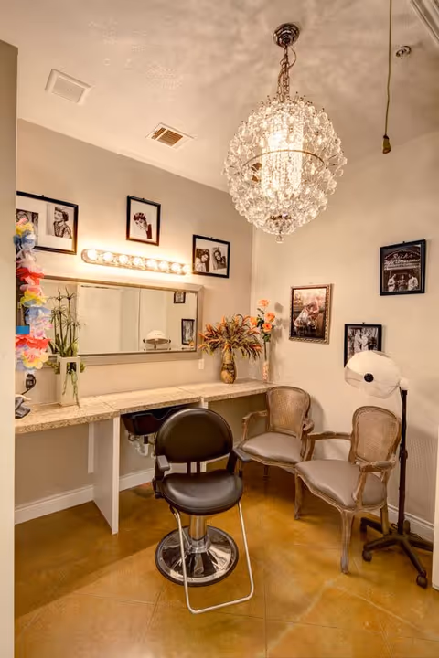 Interior room with a vanity counter and mirror, a black salon chair, two cushioned wooden chairs, a standing hair dryer, and framed black and white photos on the walls. A decorative chandelier hangs from the ceiling.