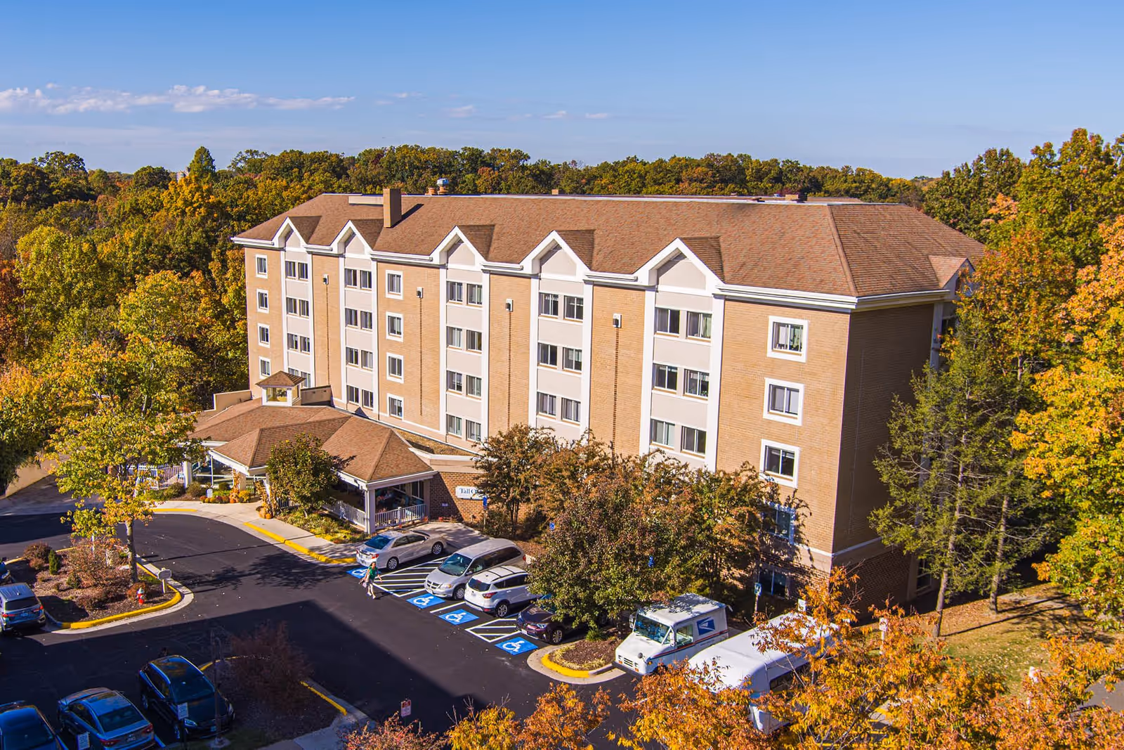 A large multi-story assisted living facility building surrounded by trees with autumn foliage. The building has a brown roof and beige walls with white trim around the windows. There is a parking lot in front with several cars and a USPS mail truck parked. The sky is clear and blue.