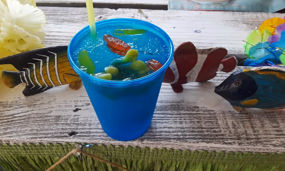 A blue plastic cup filled with a blue-colored drink and gummy worms on top, placed on a rustic wooden surface decorated with colorful artificial fish and flowers.