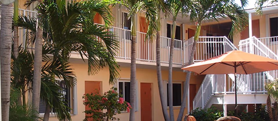 Courtyard of a two-story tropical apartment building with palm trees, balconies, and an orange patio umbrella.