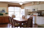A bright kitchen and dining area with a wooden table surrounded by four cushioned chairs. The kitchen features white cabinets, a microwave, a refrigerator, and various kitchen appliances on the countertop. A large window with a valance allows natural light to fill the room. There is a wooden sideboard with decorative items and plates displayed above the cabinets.