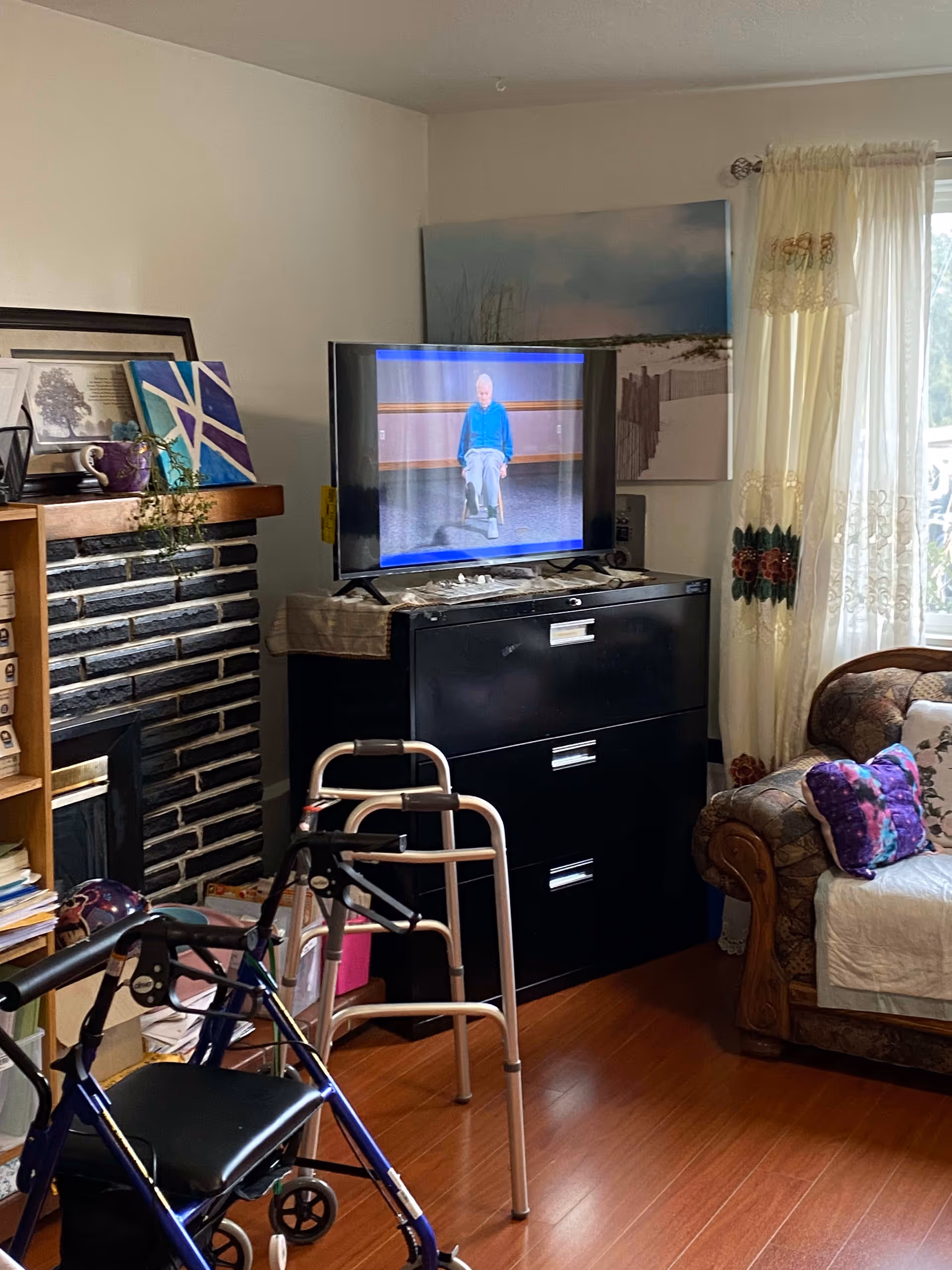 A cozy living room area with a patterned sofa adorned with colorful pillows and a white throw. A black filing cabinet holds a flat-screen TV displaying an elderly person seated on a chair. In front of the cabinet, there are two walkers, one blue and one silver. To the left, there is a fireplace with a wooden mantel decorated with framed pictures, a purple mug, and a small plant. Light-colored curtains with floral embroidery cover a window, allowing natural light to brighten the room.