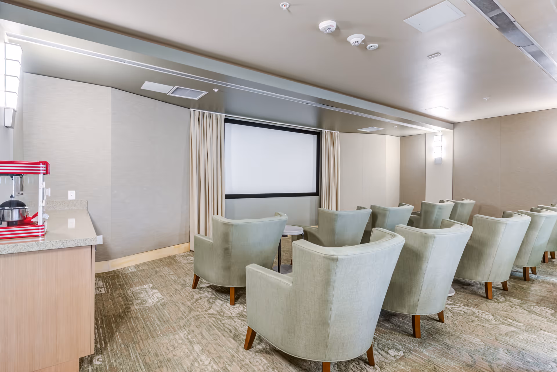 A small theater room with two rows of light green upholstered armchairs facing a blank projector screen. To the left, there is a countertop with a red and white popcorn machine. The room has beige walls, carpeted floor, and modern wall-mounted lights.
