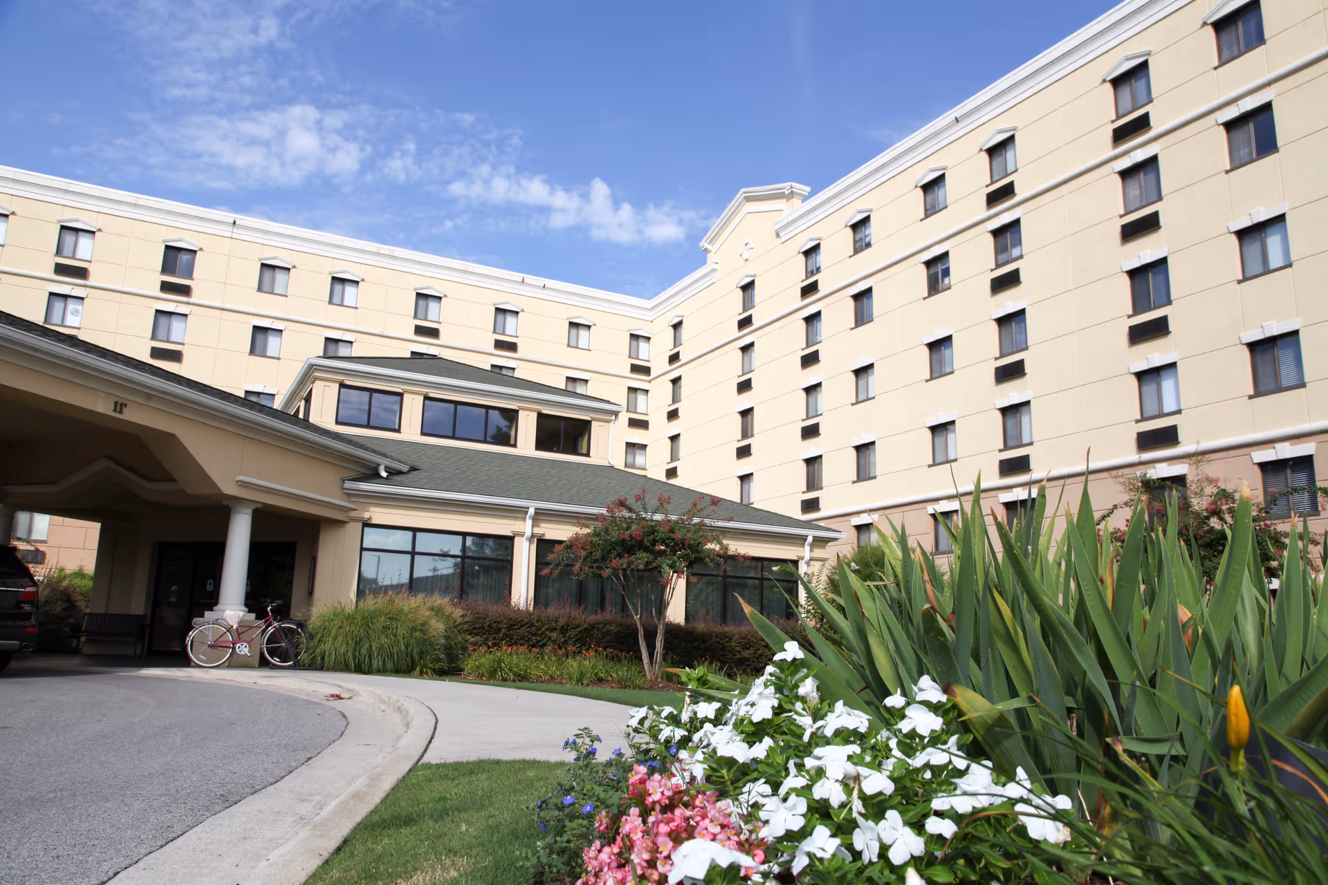Exterior view of a multi-story beige building with numerous windows, a covered entrance with white columns, and a driveway. In the foreground, there are green plants and colorful flowers under a partly cloudy blue sky.
