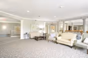 A spacious and well-lit living room area with light-colored walls and carpet. The room features several upholstered chairs and sofas arranged around a coffee table. Decorative columns and artwork adorn the walls, and there is a reception or service desk area in the background.