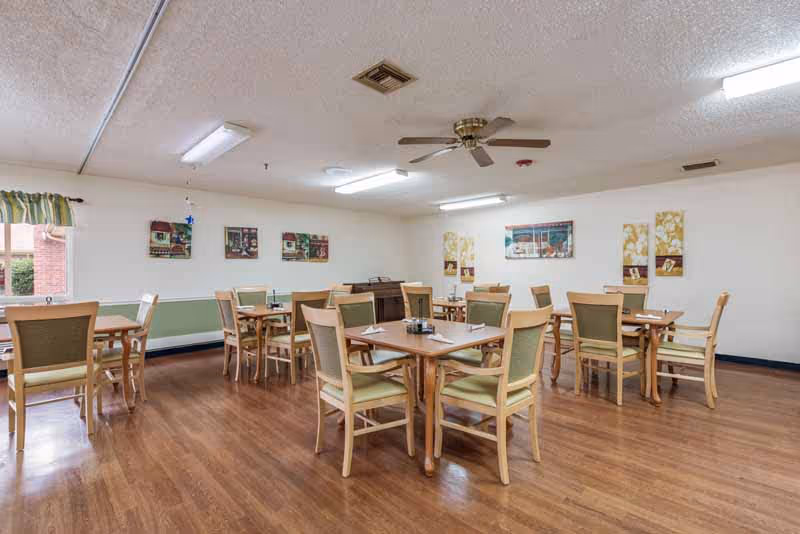 A dining room in a senior living facility with several wooden tables and chairs arranged neatly on a wooden floor. The walls are decorated with framed pictures and there is a ceiling fan and fluorescent lights on the ceiling. A window with a green valance is visible on the left side.