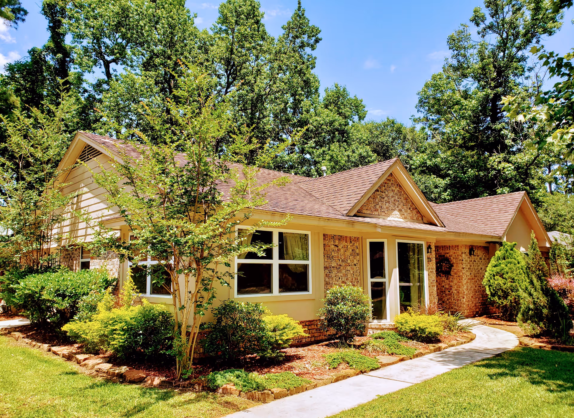 Exterior view of a single-story brick and siding building surrounded by green trees and shrubs under a clear blue sky. A curved concrete pathway leads to the entrance of the building.