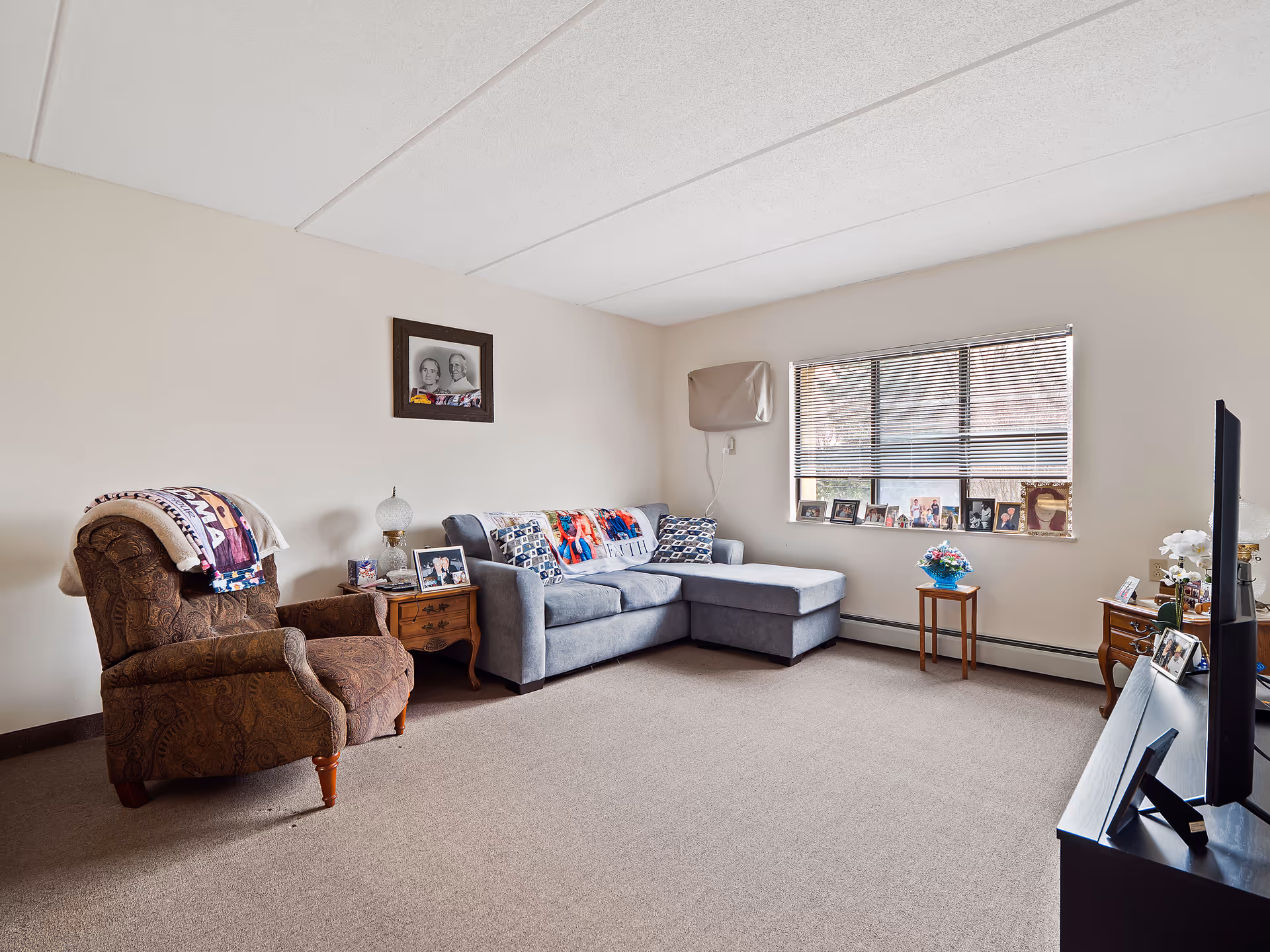 A cozy living room with a brown patterned armchair draped with blankets, a small wooden side table with a lamp and framed photos, a gray sectional sofa with colorful pillows and a throw blanket, a window with blinds and several framed photos on the windowsill, a small wooden table with a flower arrangement, and a TV on a black stand with more framed photos.