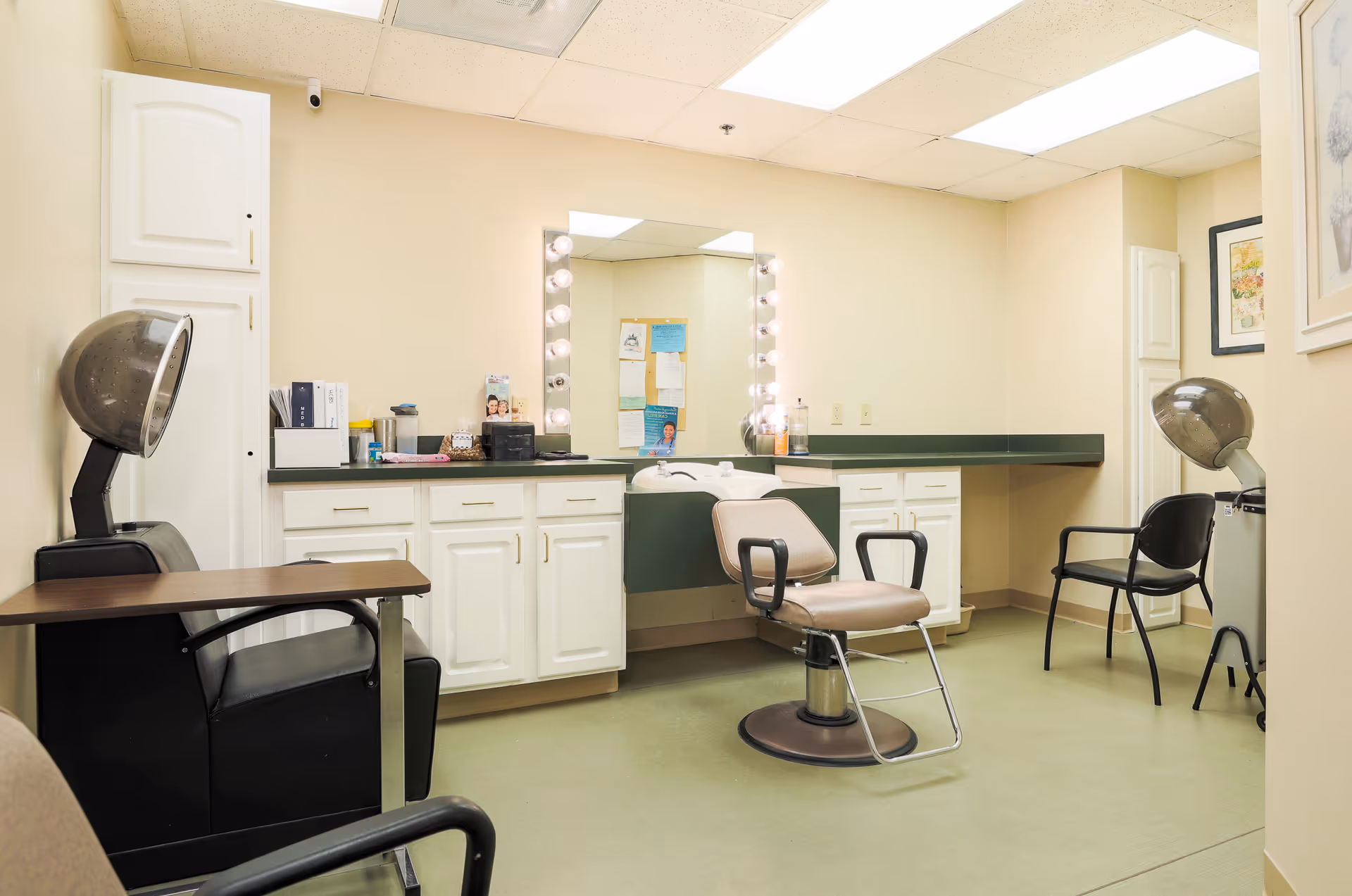 Interior view of a hair salon area in an assisted living facility with salon chairs, hair dryers, a large mirror with lights, and white cabinets along the wall.