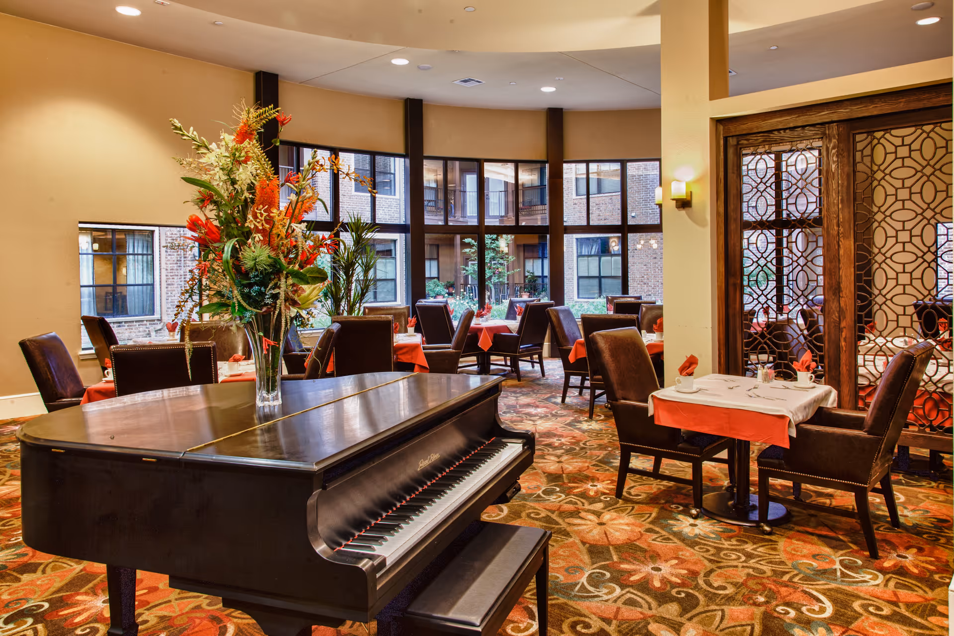 Interior view of a dining room in a senior living facility with a grand piano in the foreground decorated with a large floral arrangement. The room has multiple tables set with white tablecloths and red napkins, surrounded by dark upholstered chairs. Large windows provide a view of an outdoor courtyard area.