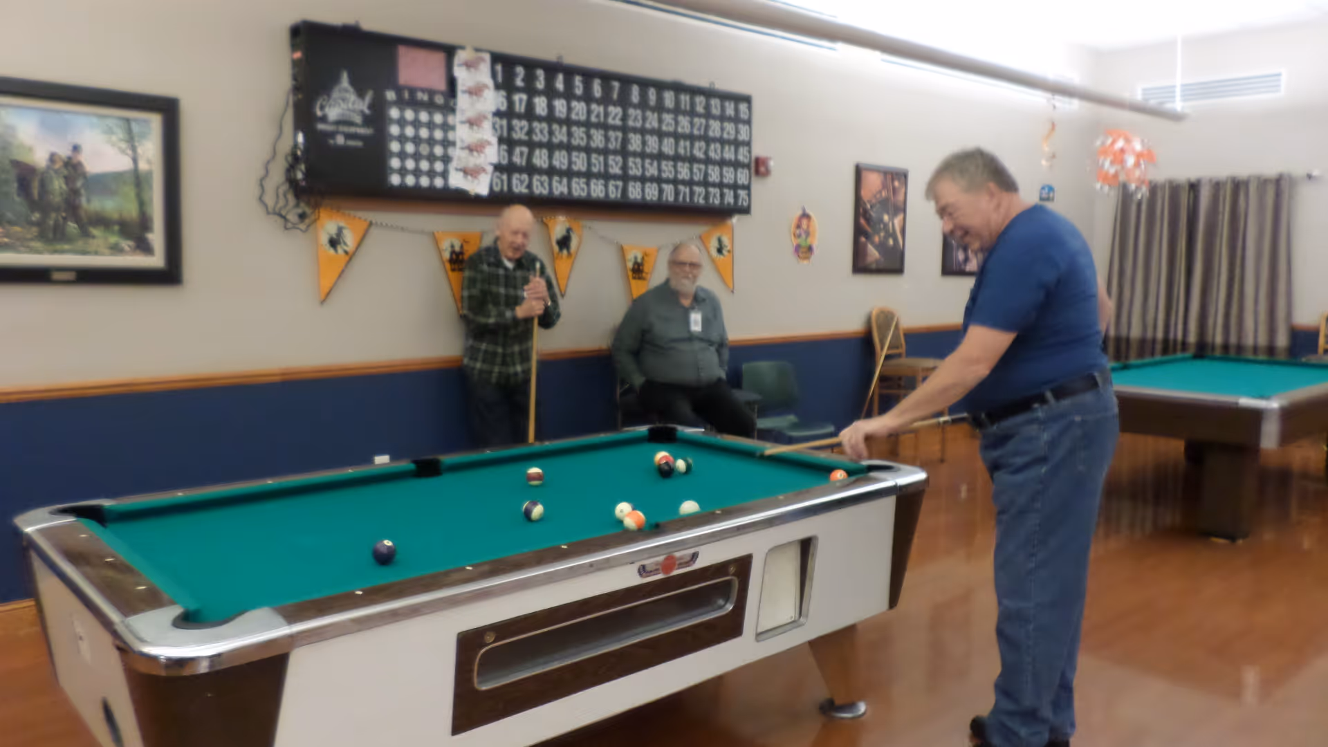 Three elderly men in a recreational room at Norfolk Veterans' Home. One man is playing pool while the other two watch and smile. The room has two pool tables, a bingo board on the wall, framed pictures, and some decorations.