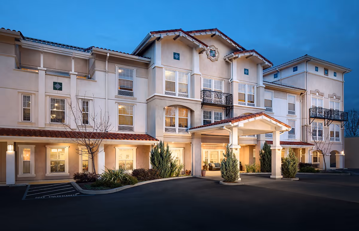 Exterior view of a three-story senior living facility building at dusk with warm lights glowing from the windows. The building features a covered entrance with columns, decorative balconies, and landscaped shrubs along the front. The sky is clear and blue, and the parking area is visible in front of the building.