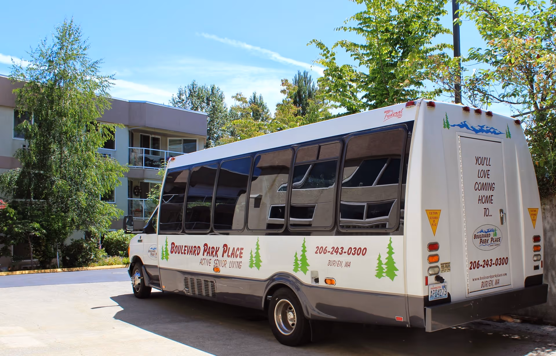 A white shuttle bus parked outside a senior living facility building. The bus has the text 'Boulevard Park Place Active Senior Living' along with contact information and a slogan 'You'll love coming home to...' on the back. The building and surrounding trees are visible in the background under a clear blue sky.