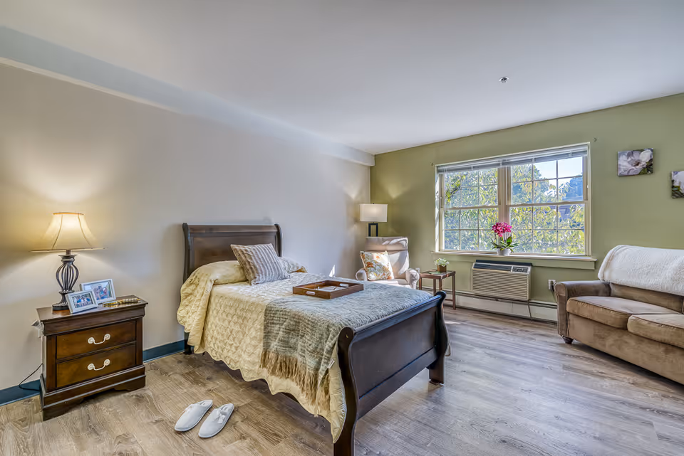A cozy bedroom in a senior living facility featuring a wooden bed with beige bedding and pillows, a wooden nightstand with a lamp and framed photos, a pair of white slippers on the wooden floor, a comfortable armchair with a floral pillow, a small side table with a plant, a large window with a view of greenery outside, and a brown sofa with a white throw blanket.