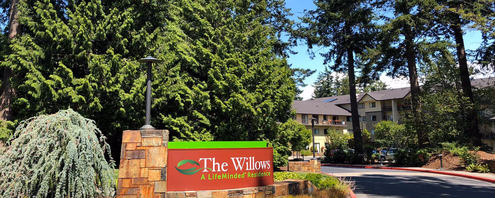 Entrance sign for The Willows, a LifeMinded Residence, surrounded by trees and landscaping with a multi-story building in the background under a clear blue sky.