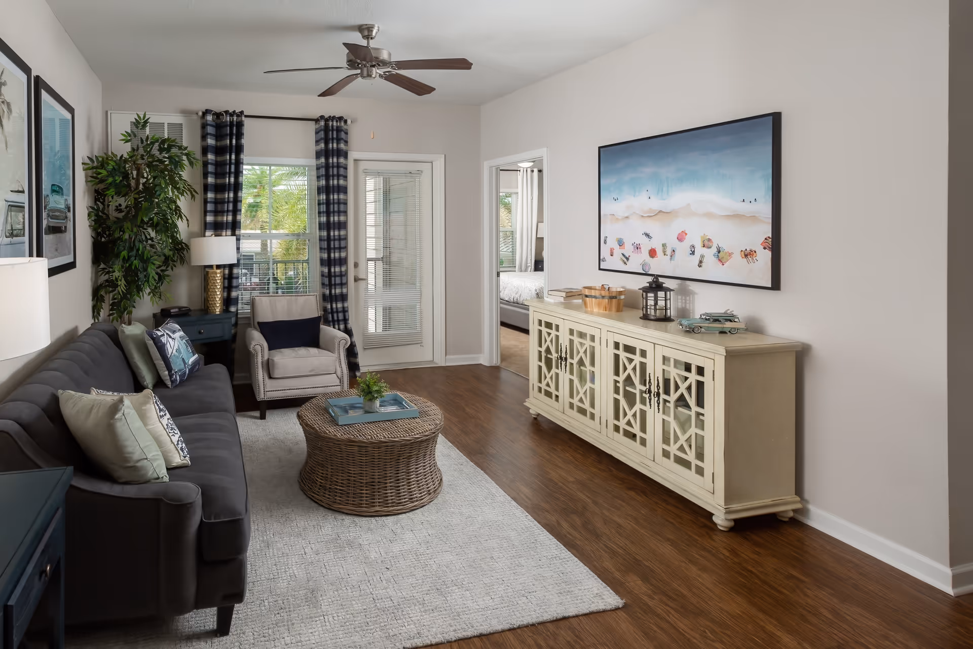 A cozy living room with a dark gray sofa adorned with multiple pillows, a beige armchair, and a round wicker coffee table on a light gray rug. There is a ceiling fan above, a large window with plaid curtains, and a glass door leading outside. On the right wall, a cream-colored cabinet with decorative lattice doors holds books, a lantern, and a model car, with a large beach-themed painting hanging above it. The room has wooden flooring and neutral-colored walls.