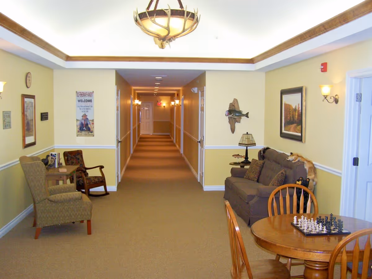 A long hallway in a senior living facility with beige walls and carpet. On the left side, there are two chairs and a small table with books. On the right side, there is a brown couch with pillows, a side table with a lamp, a mounted fish decoration, and a framed painting of a forest path. In the foreground, there is a round wooden table with a chessboard and four chairs. The ceiling has a decorative light fixture resembling antlers.