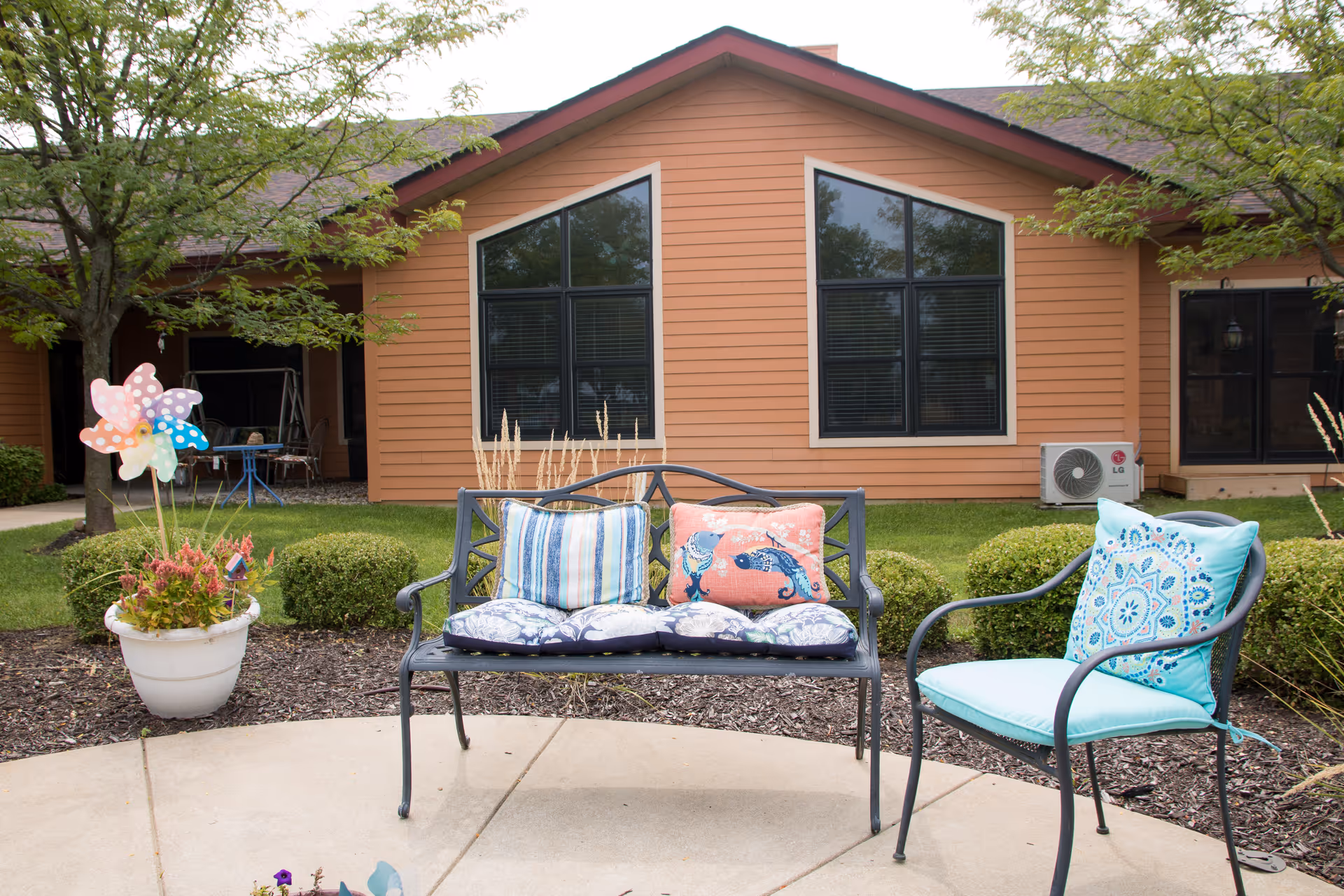 Outdoor patio area in front of a single-story building with a bench and chair with colorful cushions, potted plants, and decorative pinwheels.