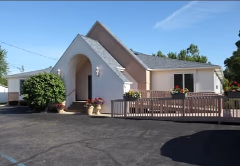 Exterior view of a single-story building with a steeply pitched roof and an arched entrance. The building has light-colored walls and a ramp with railings leading to the entrance. There are potted plants and flowers near the entrance and along the ramp. The area in front of the building is paved, and there are trees and a clear blue sky in the background.