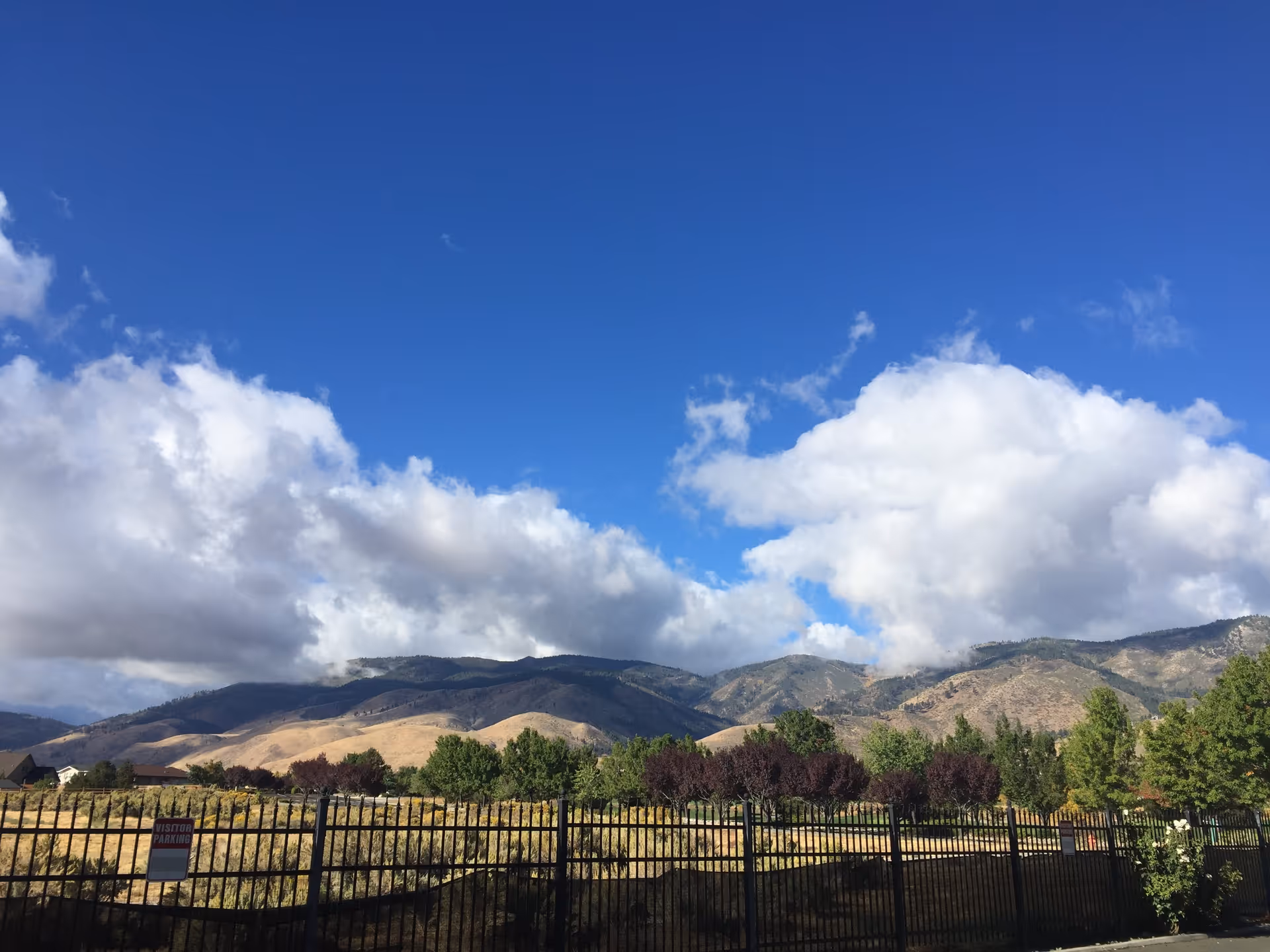 Scenic view of a mountain range under a blue sky with scattered clouds, seen over a black metal fence with a visitor parking sign. Trees and shrubs are visible in the foreground.