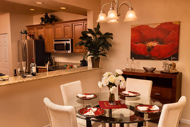 Dining area with a round glass table set for four in front of an open kitchen and a large red flower painting on the wall.