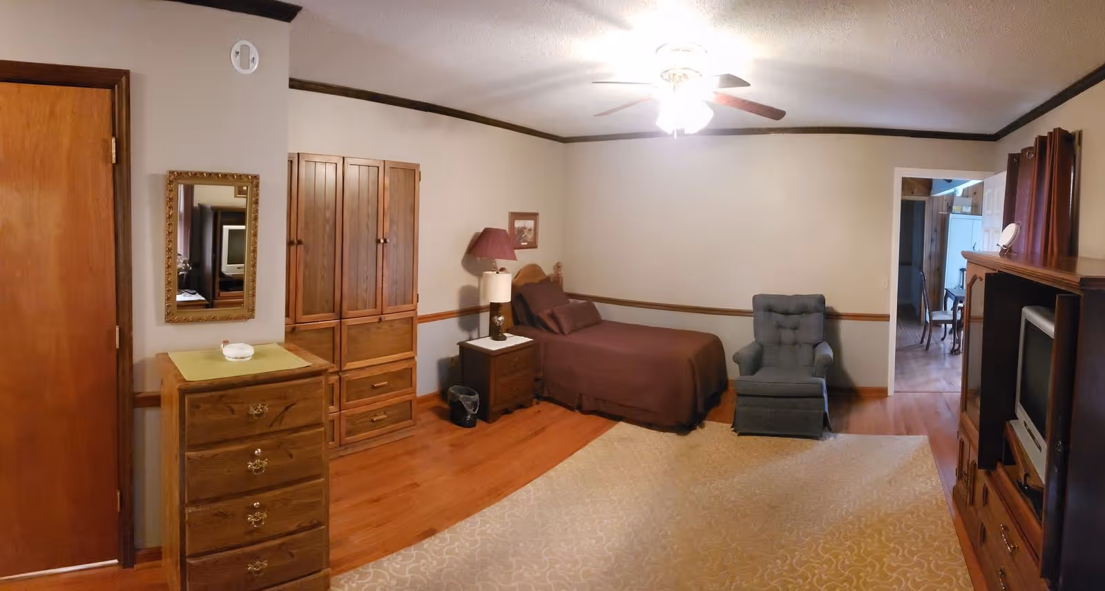 A cozy bedroom with wooden furniture including a dresser, armoire, and nightstand. The bed is made with brown bedding and is positioned against a beige wall. There is a blue upholstered armchair next to the bed. A ceiling fan with lights is on the ceiling. The room has hardwood floors with a light-colored patterned area rug. A TV is placed in a wooden entertainment center opposite the bed. A doorway leads to another room with visible chairs and a white cabinet.