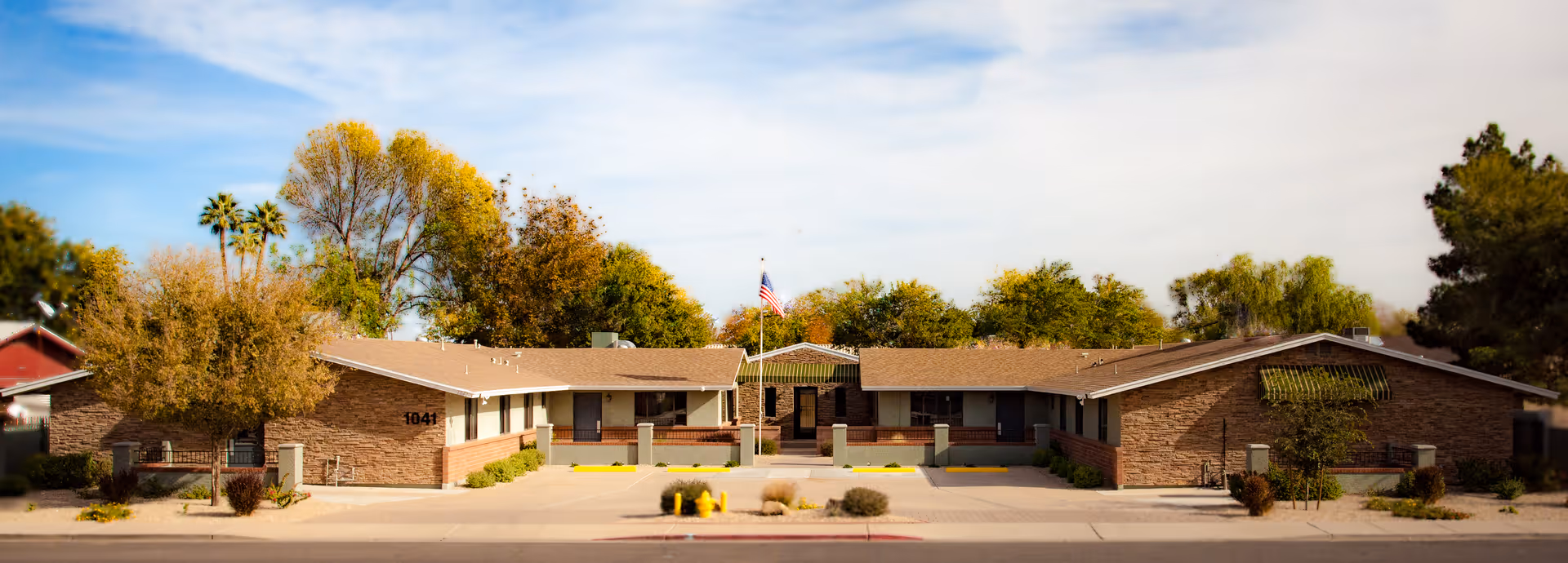 Exterior front view of a single-story brick building with a central entrance, surrounded by trees and bushes under a partly cloudy sky. The building has a flagpole with an American flag in front and the number 1041 displayed on the left side.