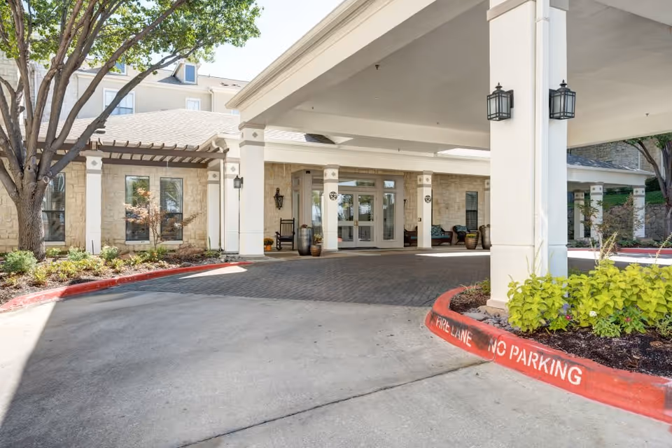 Entrance area of Ridgmar Place facility showing a covered driveway with columns, outdoor seating, landscaped plants, and a tree. The building exterior features stone walls and multiple windows.