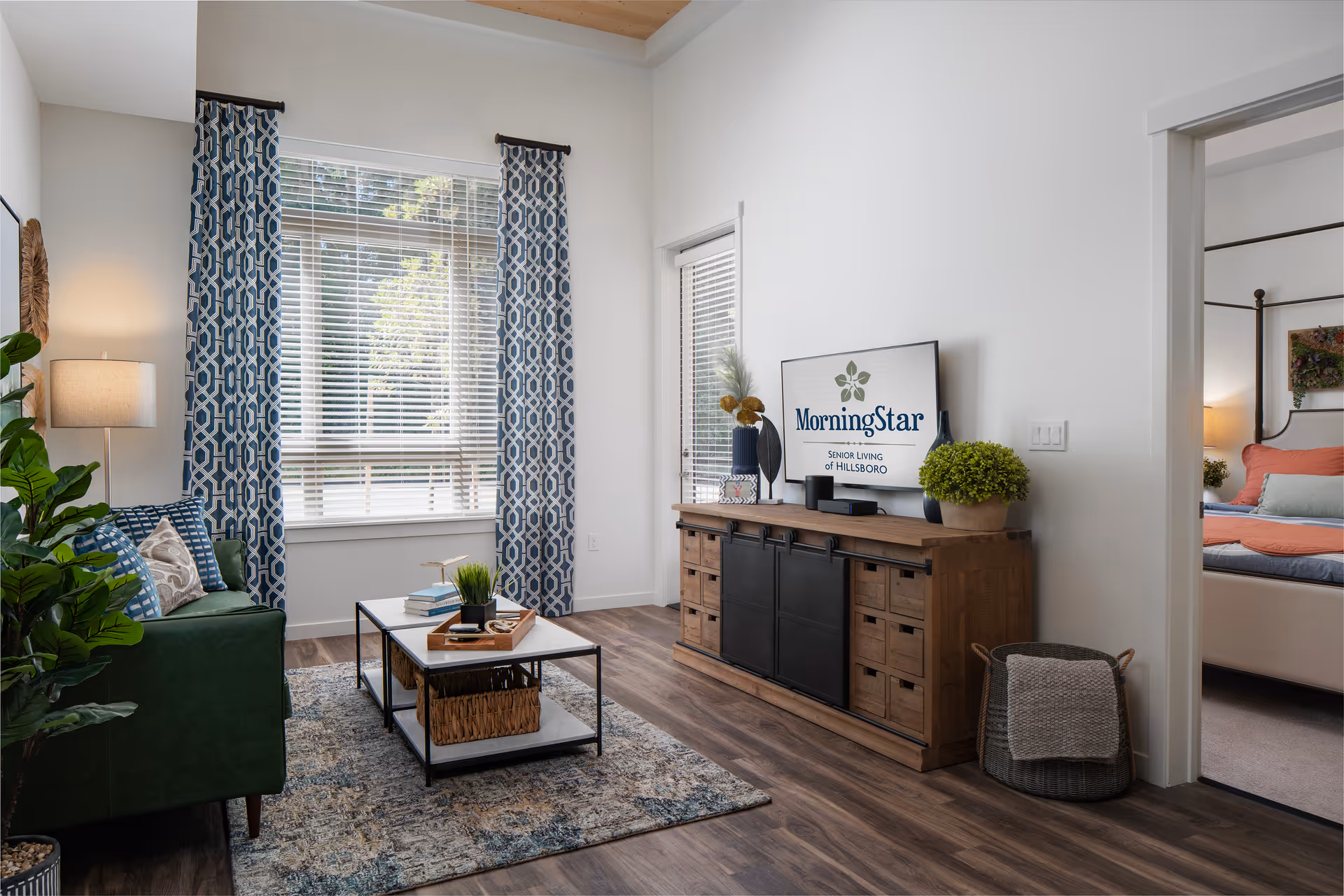 A bright and cozy living room with a green sofa adorned with patterned pillows, a floor lamp, and a coffee table with books and a plant. The room features large windows with blue and white patterned curtains, wooden flooring, and a wooden media console with decorative items and a TV displaying the MorningStar Senior Living of Hillsboro logo. An adjacent bedroom with a bed and colorful bedding is visible through an open doorway.