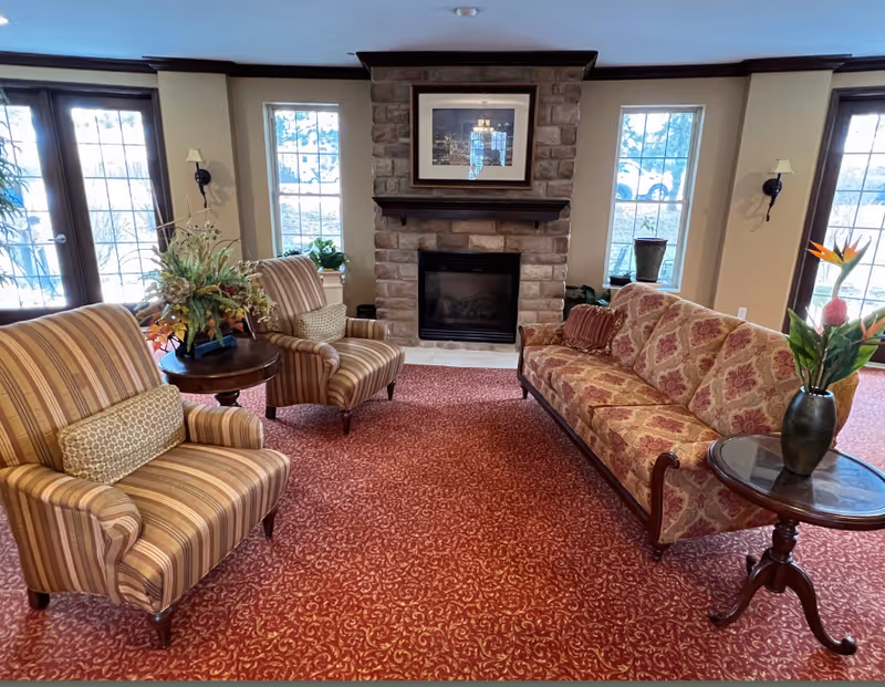 A cozy living room area with a patterned red carpet, a stone fireplace in the center, and three large windows letting in natural light. The room features a floral-patterned sofa, two striped armchairs with cushions, and two wooden side tables with decorative plants and flowers.