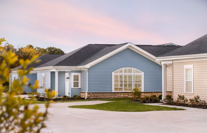 Exterior view of a single-story residential building with blue and beige siding, a large arched window, and a paved driveway surrounded by some greenery and small bushes under a pinkish sky at sunset.