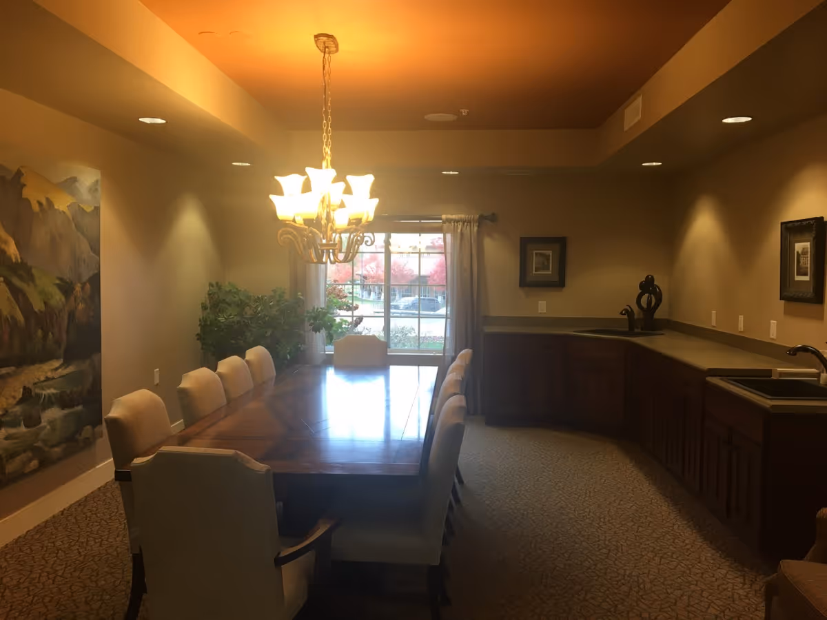 A dining room with a long wooden table surrounded by upholstered chairs. A chandelier with multiple lights hangs above the table. There is a large window with curtains at the far end, letting in natural light. The room has beige walls, carpeted floor, and a countertop with a sink and cabinets along one side. A large painting of a mountain landscape is on one wall, and there are framed pictures on another wall.