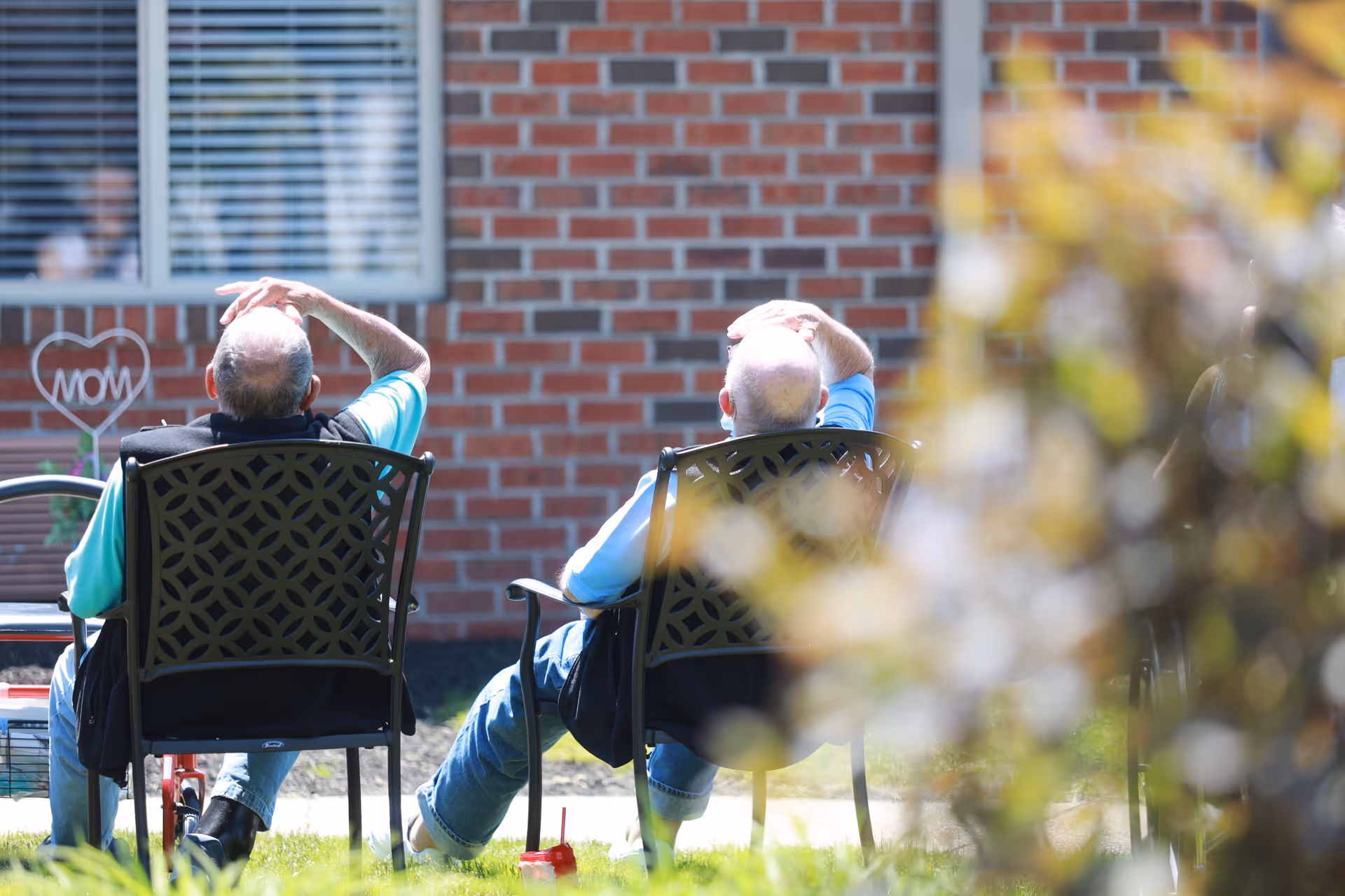 Two elderly men sitting on outdoor chairs facing a brick building, both shielding their eyes from the sun with one hand. There is a heart-shaped decoration with the word 'MOM' visible near the window in the background.