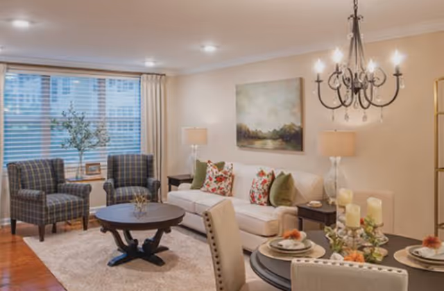 A cozy living room with a white sofa adorned with floral and green pillows, two plaid armchairs, a round wooden coffee table on a beige rug, and a dining table set with plates and candles. The room features a large window with blinds and curtains, two table lamps, a chandelier, and a landscape painting on the wall.