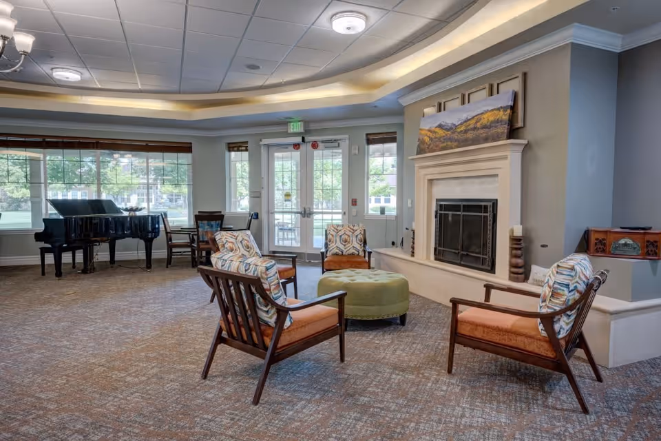 A spacious living room area with four wooden chairs with patterned cushions arranged around a green round ottoman. There is a fireplace with a landscape painting above it on the right wall. Large windows and glass doors let in natural light, and a black grand piano is positioned near the windows on the left side of the room.