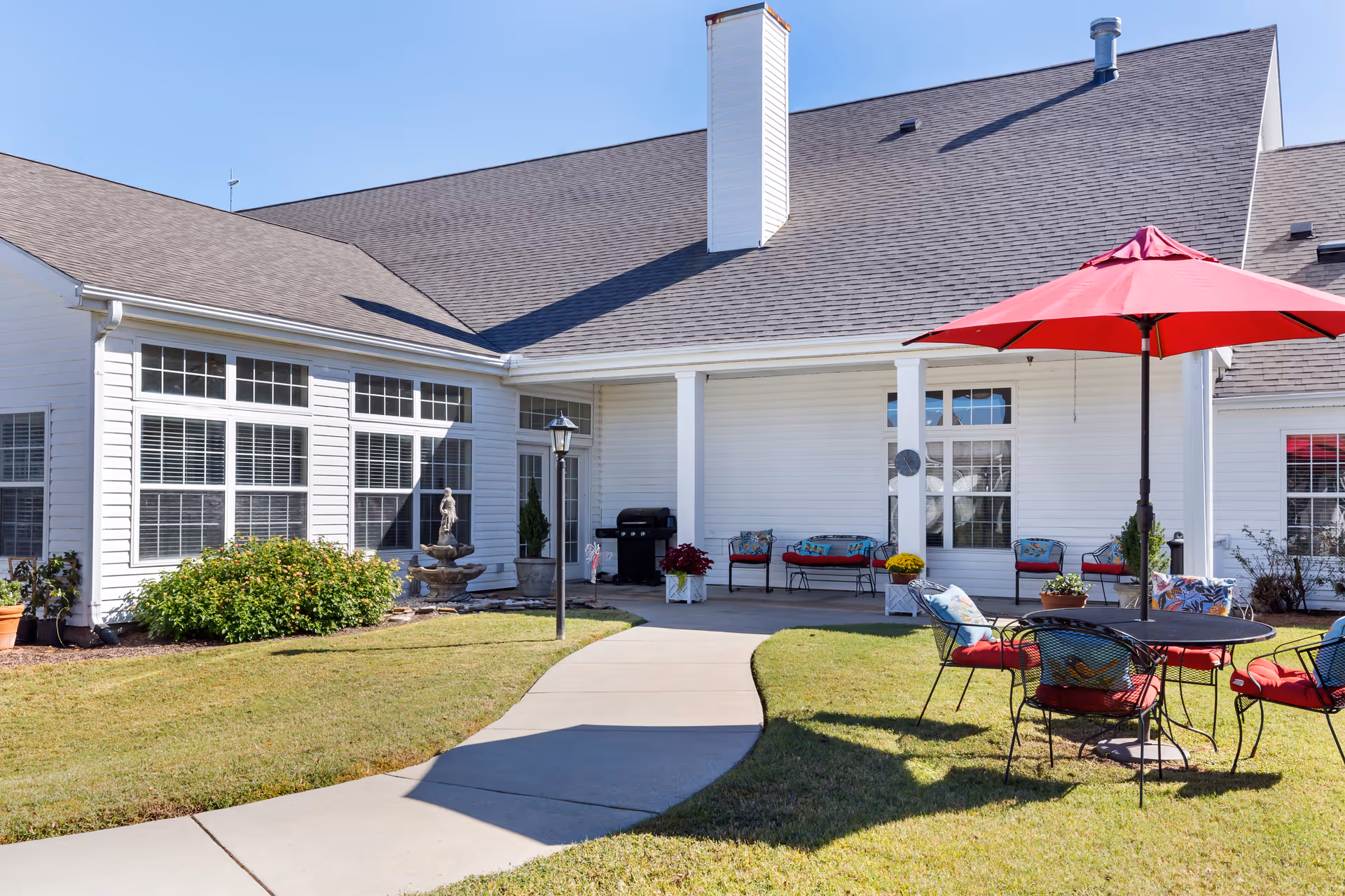Outdoor courtyard and entrance of a white senior living building with patio furniture, a red umbrella, and a curved walkway.