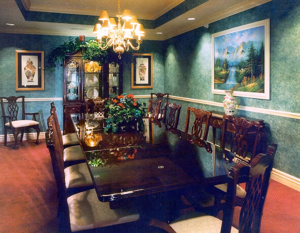 A formal dining room with a long polished wooden table surrounded by cushioned wooden chairs. The walls are green with white trim and decorated with framed artwork and a large landscape painting. A chandelier with multiple lights hangs above the table. There is a wooden china cabinet displaying plates and glassware, and a sideboard with a decorative vase. A floral centerpiece adorns the dining table.