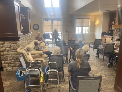 A group of elderly people seated in a common room facing two musicians playing guitars near a window. The room has stone walls, a fireplace, and several walkers are visible in the foreground.