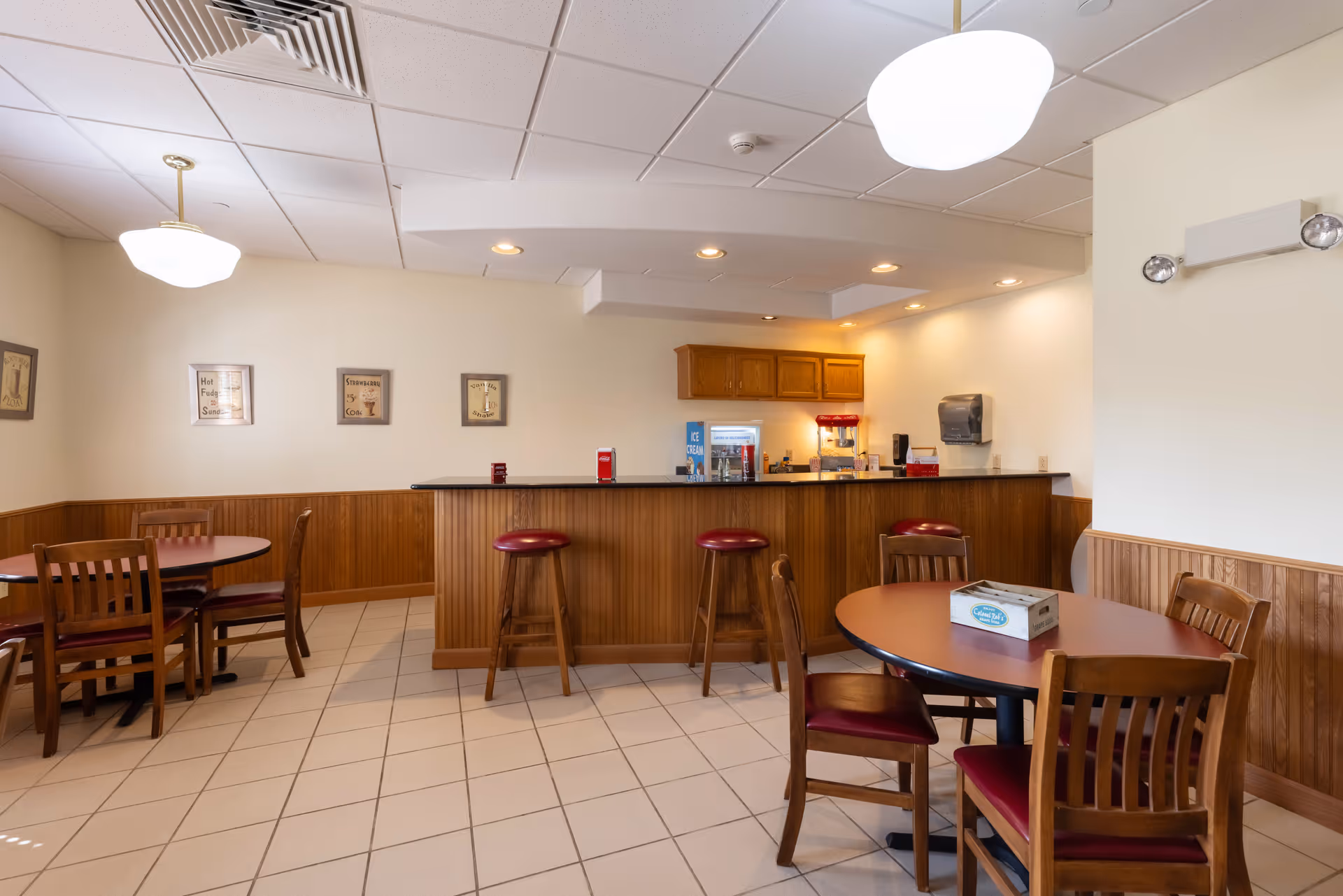 A dining area in Prairie Hills at Independence featuring round wooden tables with chairs that have red cushioned seats. There is a wooden counter with three bar stools in front of a small kitchen or serving area. The walls are light-colored with wood paneling on the lower half, and there are framed pictures hanging on the wall. The ceiling has recessed lighting and two hanging light fixtures.